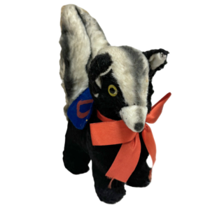 Wee Stinker Collegiate Skunk. The image shows a small stuffed toy wearing a blue felt badge with an orange letter "D" on it and an orange ribbon around its neck. The toy has a black body with a white strip along its back and head, yellow eyes, and a white snout, The tail is raised, and the fur looks worn due to its age.