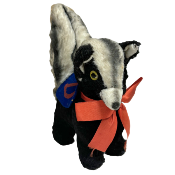 Wee Stinker Collegiate Skunk. The image shows a small stuffed toy wearing a blue felt badge with an orange letter "D" on it and an orange ribbon around its neck. The toy has a black body with a white strip along its back and head, yellow eyes, and a white snout, The tail is raised, and the fur looks worn due to its age.